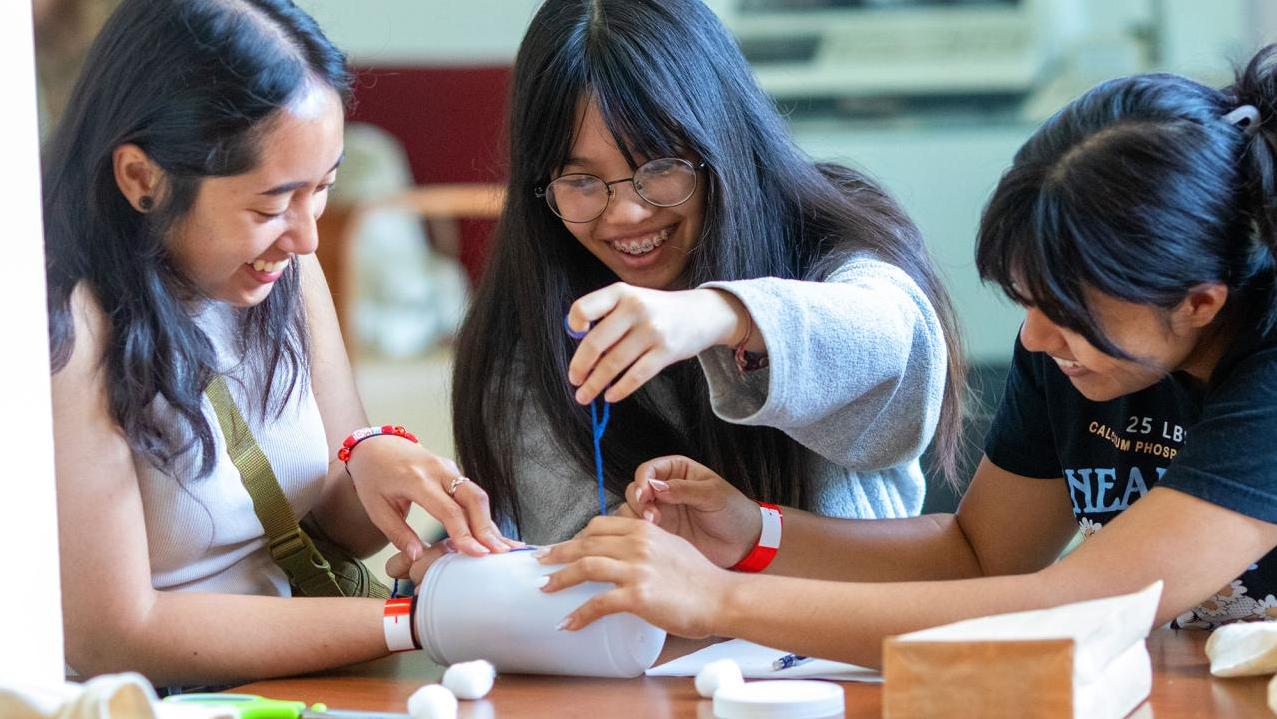 Three students smiling and assembling a small craft project with tape in a classroom