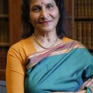 A woman in a green and orange saree smiles warmly, standing in a library setting.